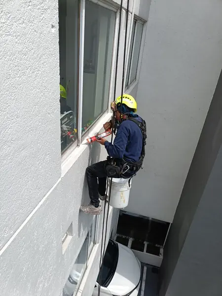 Trabajador con casco y arnés sostiene una pistola de sellador contra la junta inferior de una ventana.