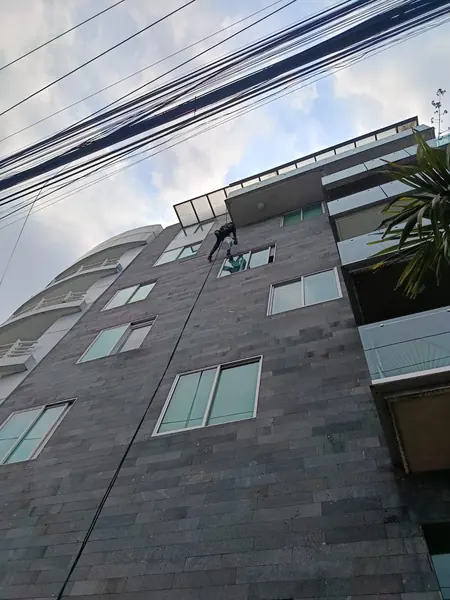 Fachada gris de varios niveles vista desde la calle, con un trabajador suspendido cerca de las ventanas superiores.
