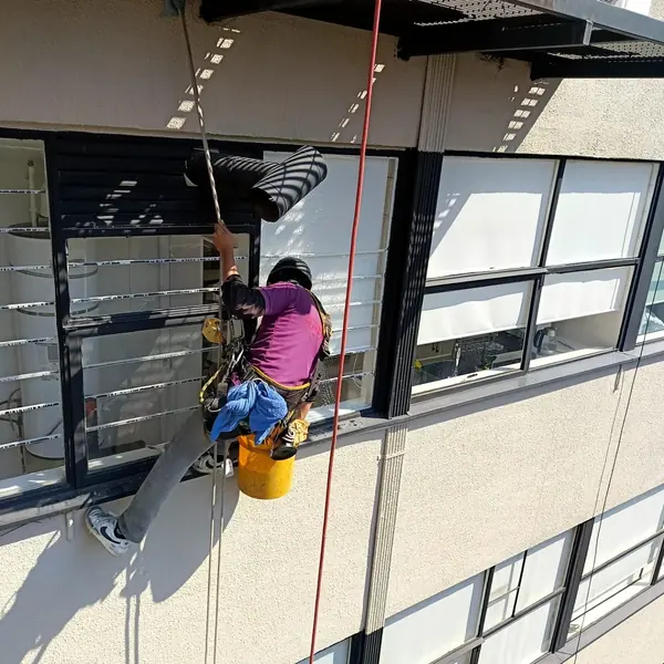 Trabajador con casco negro y cubeta amarilla suspendido junto a un ventanal con espuma y escurrimientos.