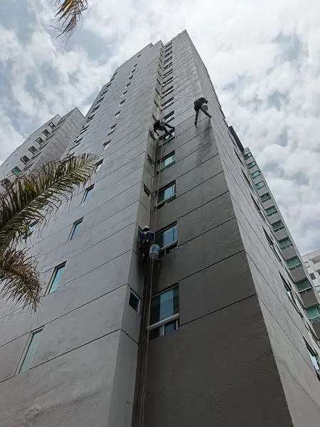 Tres trabajadores suspendidos a diferentes alturas en una fachada gris, vistos desde la base del edificio.
