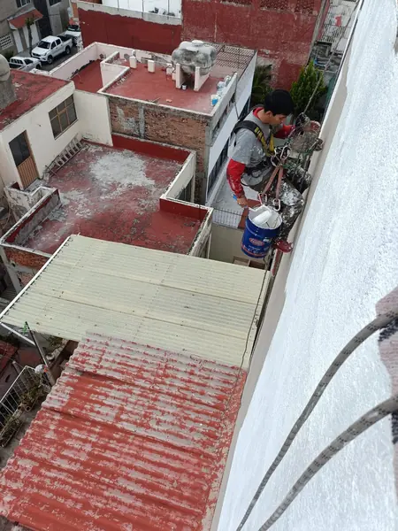 Trabajador suspendido junto a un muro blanco sostiene un rodillo cargado de material blanco sobre una cubeta azul.
