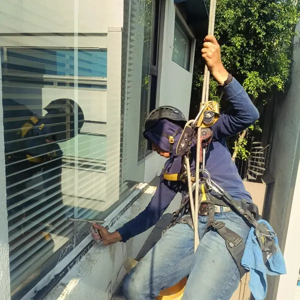 Trabajador con casco negro utiliza un jalador en T sobre la parte inferior de un vidrio con espuma mientras se sostiene de una cuerda.