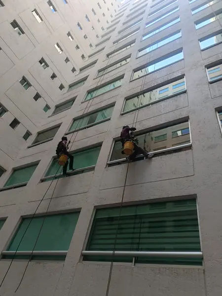 Dos trabajadores suspendidos junto a ventanas de un patio alto y estrecho, con cubetas amarillas y escurrimientos sobre los cristales.
