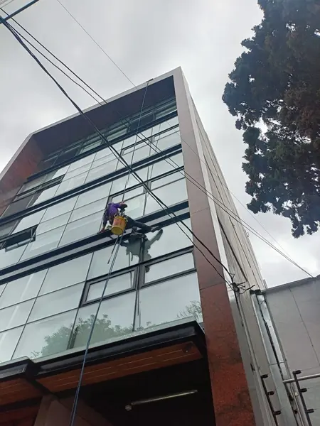 Trabajador suspendido con cubeta amarilla y utensilio de mango largo frente a una fachada de vidrio vista desde abajo.