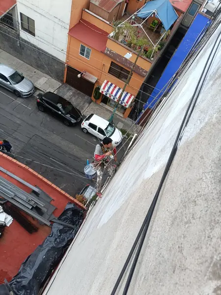 Trabajador suspendido manipula sus cuerdas junto a un muro con zonas blancas y grises; lleva una cubeta azul con material blanco.