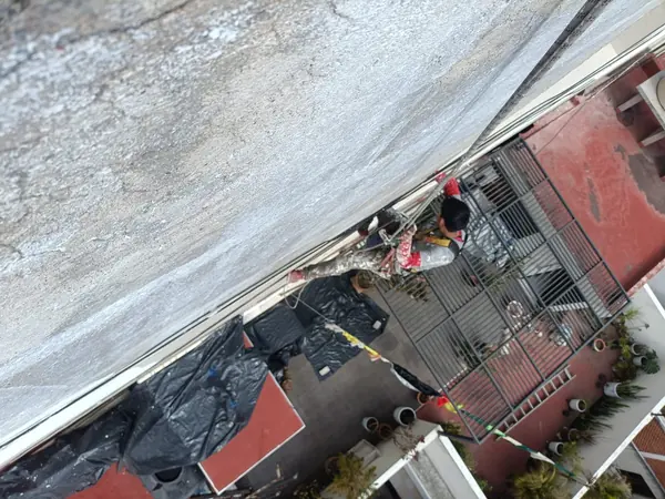 Vista desde arriba de un trabajador suspendido junto al borde de un muro blanco y gris, con protección plástica negra sobre el área inferior.