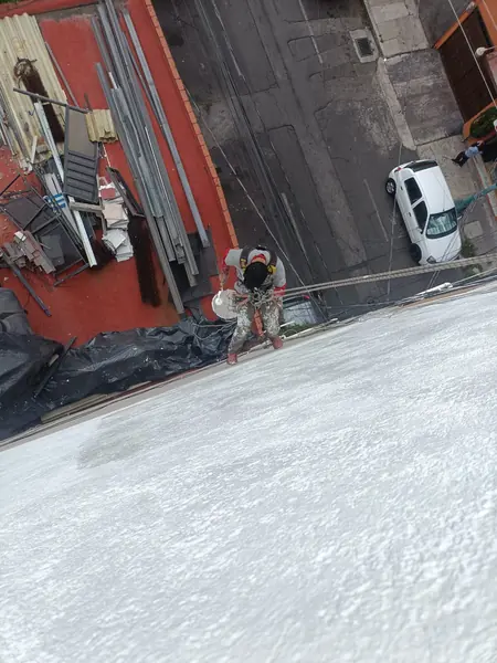 Vista cenital de un trabajador suspendido que sostiene un rodillo blanco sobre una cubeta junto a un muro con recubrimiento.