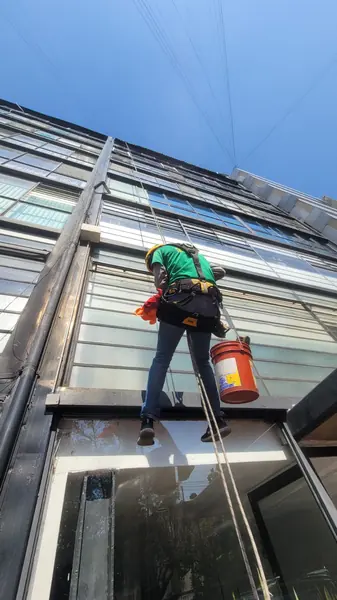 Trabajador de espaldas, con camisa verde, arnés y cubeta naranja, suspendido frente a un ventanal de franjas horizontales.