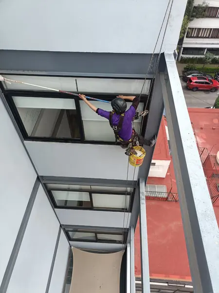 Trabajador suspendido con cubeta amarilla sostiene un utensilio de mango largo sobre un ventanal, visto desde la azotea.