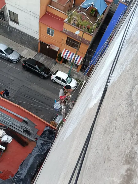 Trabajador suspendido ajusta su posición mediante cuerdas junto a un muro blanco y gris, con una cubeta azul a su costado.