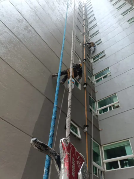 Cuerdas y un dispositivo rojo de descenso en primer plano, con trabajadores suspendidos junto a ventanas al fondo.