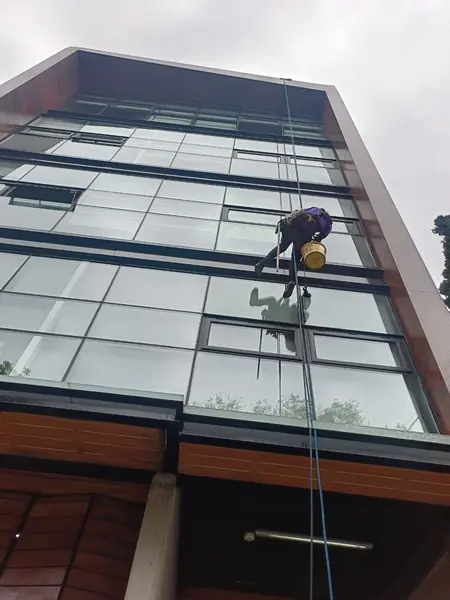 Trabajador suspendido frente a una fachada acristalada sostiene un utensilio de limpieza de mango largo y una cubeta amarilla.