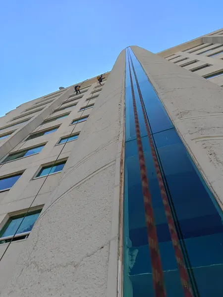 Fachada con una franja central de vidrio azul, cuerdas que recorren su altura y trabajadores pequeños cerca de la parte superior.