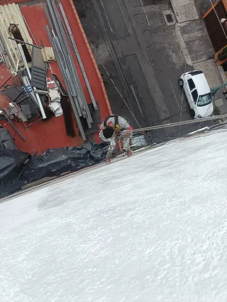 Vista desde arriba de un trabajador suspendido junto a un muro blanco, con cuerdas y cubiertas de protección en el nivel inferior.
