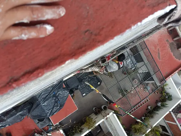Trabajador suspendido junto a una fachada, visto desde un borde rojo de azotea, con un patio y una estructura metálica debajo.