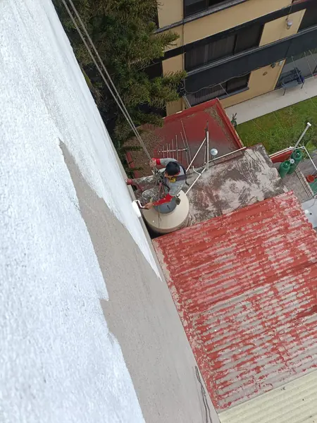 Trabajador suspendido por cuerdas pasa un rodillo blanco sobre un muro exterior, con azoteas y un tinaco visibles abajo.