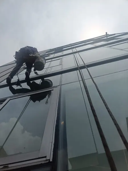 Vista desde abajo de un trabajador suspendido ante paneles de vidrio, con una cubeta y cuerdas que suben por la fachada.