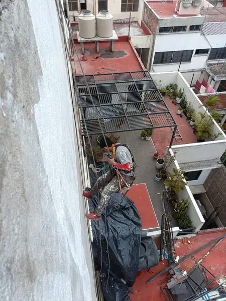 Trabajador con arnés y asiento suspendido junto a un muro parcialmente blanco, sobre patios con macetas y cubiertas negras.