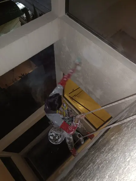 Trabajador suspendido en un patio interior extiende un atomizador hacia el muro situado sobre una ventana iluminada.