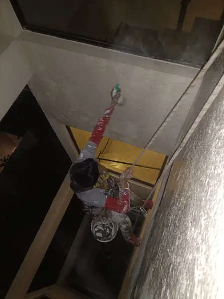 Trabajador sujeto a cuerdas apunta un atomizador de gatillo verde al muro blanco entre dos ventanas del patio interior.