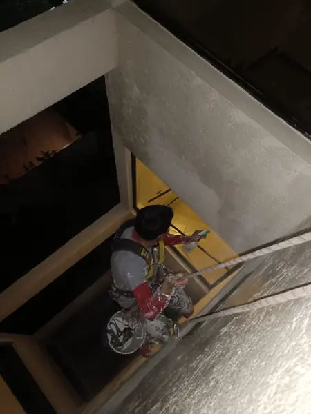 Trabajador suspendido junto a una ventana iluminada sostiene un atomizador en una mano y una cuerda en la otra.