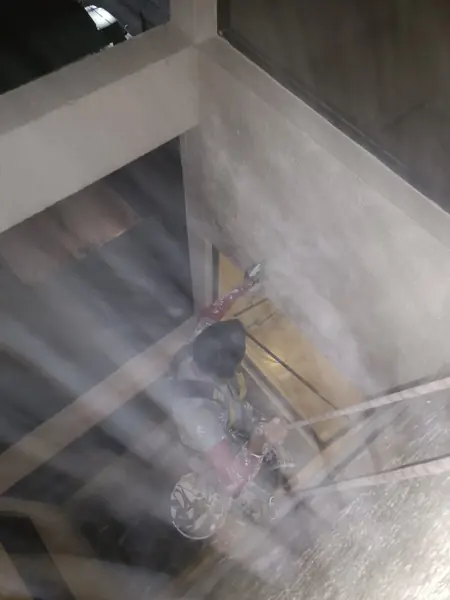 Trabajador suspendido dirige un atomizador hacia el muro entre ventanas; un velo claro cruza parte de la imagen.