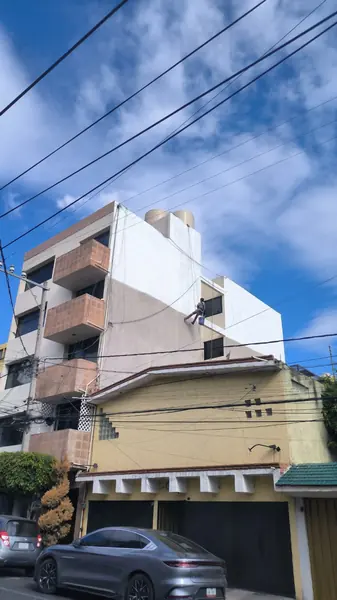 Edificio con balcones de ladrillo y muro blanco y gris, con un trabajador suspendido en la fachada y cables en primer plano.
