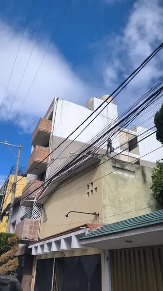 Vista lateral desde la calle de un edificio con balcones, cuerdas y un trabajador suspendido, parcialmente cruzada por cables aéreos.