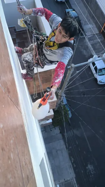 Trabajador suspendido frente a la fachada sostiene una cuerda y un rociador blanco con cabezal naranja junto al muro.