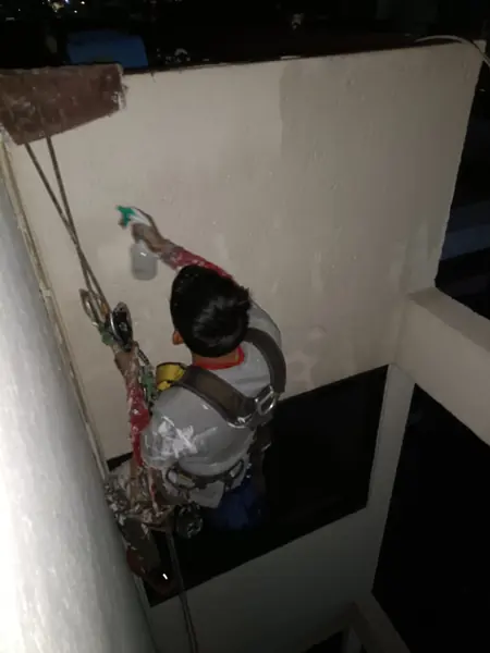Trabajador con arnés extiende un atomizador de gatillo verde hacia un muro blanco, junto a una esquina del patio interior.