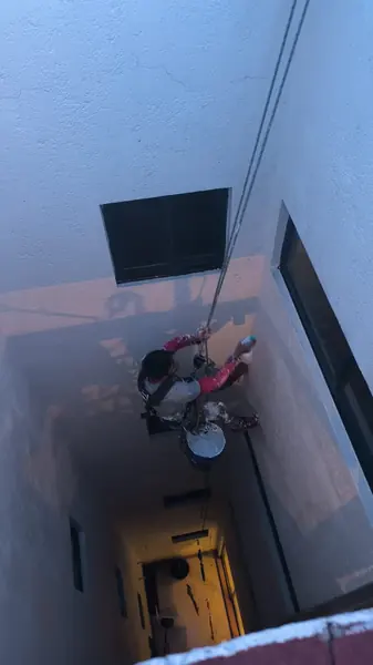 Trabajador suspendido bajo una ventana negra dirige un atomizador al muro del patio, con una cubeta colgada del asiento.