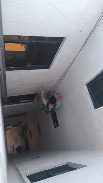 Vista desde arriba de un trabajador suspendido entre ventanas de un patio interior, con atomizador en una mano y cuerdas en la otra.