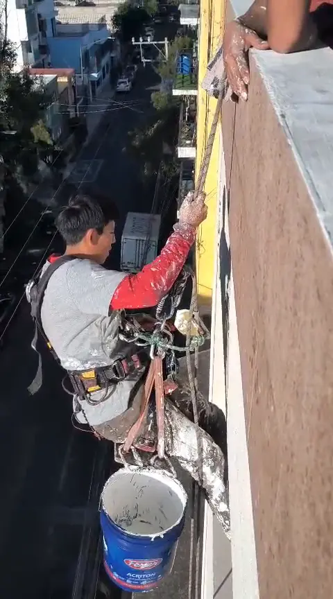 Vista cercana del operario con arnés, cuerdas, cubeta y rociador manual junto a la fachada.