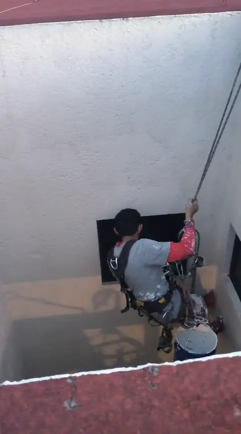 Operario con arnés y cuerdas frente a un muro blanco y una ventana oscura del patio.