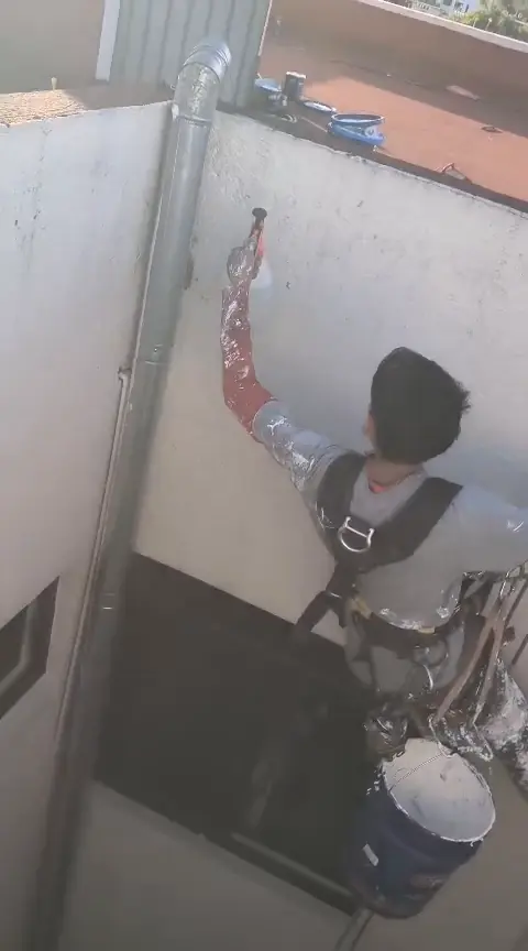 Operario con cuerdas, cubeta y rociador manual junto a una tubería vertical y un muro blanco.
